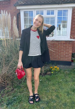 Blazer negro sobre una camiseta de rayas con un corazón rojo, combinado con pantalones cortos negros y sandalias negras. Sosteniendo un pequeño bolso rojo.
