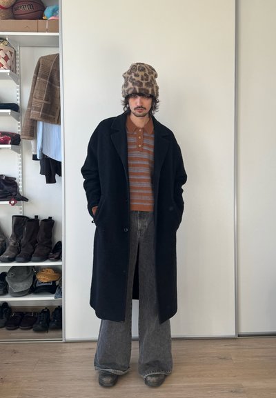 Hombre con un sombrero de estampado de leopardo, polo rayado marrón, abrigo negro, jeans anchos grises y botas, de pie en el interior cerca de un armario blanco con estantes de zapatos y ropa.