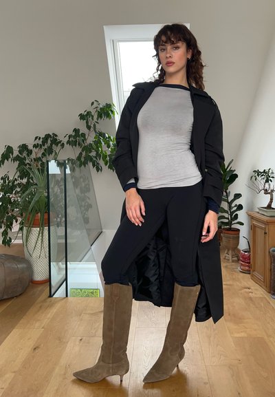 Abrigo negro sobre una camiseta gris, leggings negros y botas altas de ante marrón. Presenta tacones altos y una silueta ajustada. Entorno interior con plantas.