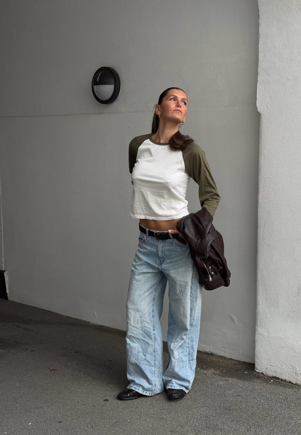 Une femme pose près d’un mur gris, tenant une veste en cuir foncé et portant un top blanc et vert, un jean bleu clair ample et des chaussures noires.