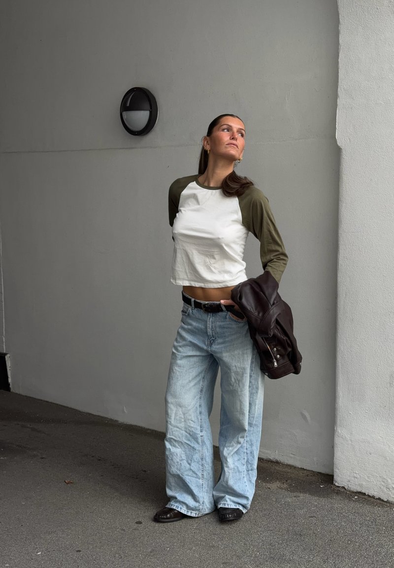 Une femme pose près d’un mur gris, tenant une veste en cuir foncé et portant un top blanc et vert, un jean bleu clair ample et des chaussures noires.