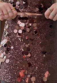 Des mains ornées de plusieurs bagues ajustant une robe brune à paillettes, agrémentée de grosses et petites décorations circulaires brillantes.