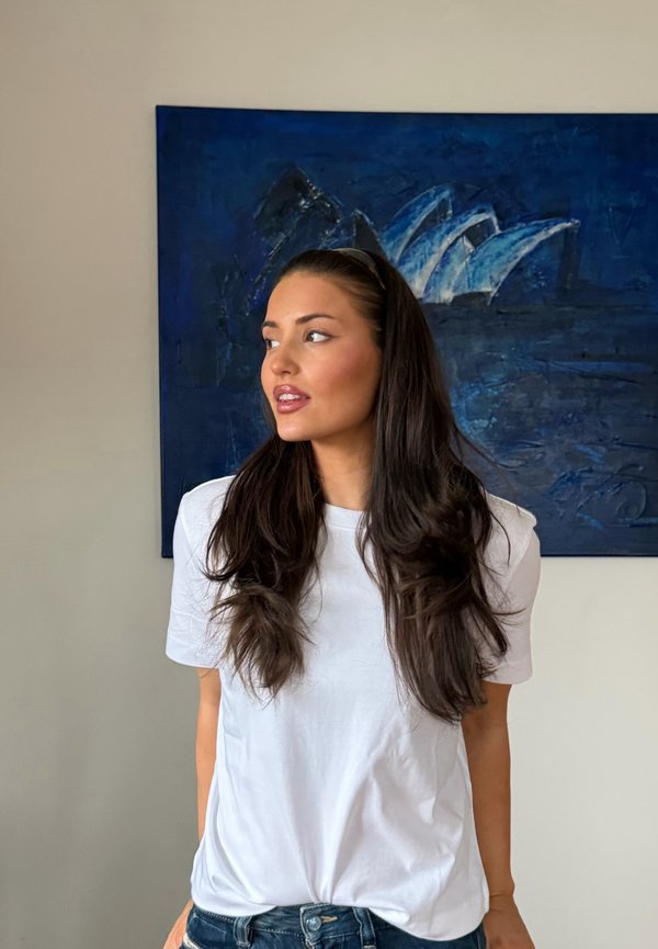 Femme aux cheveux longs et foncés, portant un T-shirt blanc et un jean, se tient de profil devant une peinture abstraite bleue sur un mur beige.