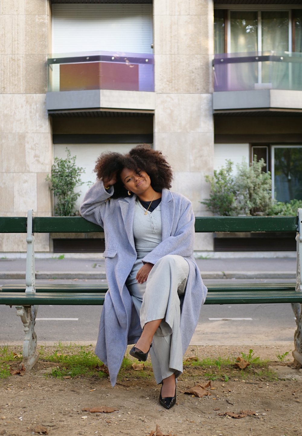 Ανοιχτό μωβ μακρύ παλτό, γκρι μακρυμάνικο μπλουζάκι, φαρδιά ανοιχτόπαντα παντελόνια, μαύρες γόβες. Καθισμένη σε ένα πράσινο παγκάκι σε αστικό περιβάλλον.
