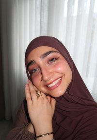 Brūns hijabs, kas sedz galvu, mīksts audums; smaidīga persona ar zilām acīm, valkājot zelta gredzenu un aproci, pret vieglām aizkariem.
