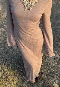 Person som bär en långärmad, ribbad mauvefärgad klänning med ett pärlhalsband, går på en torr, stenig utomhusyta.