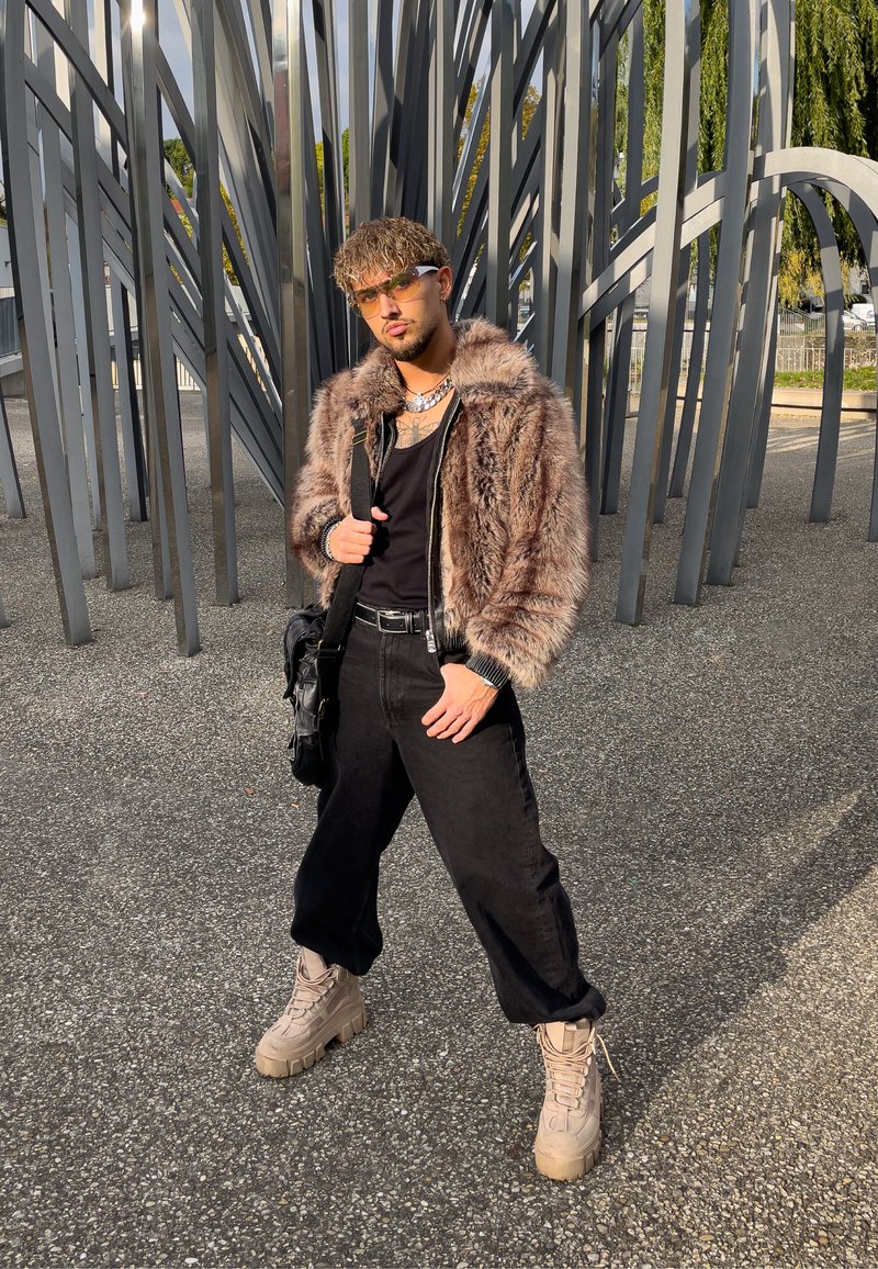 Nepbonten jas in bruintinten, zwarte tanktop, losse zwarte broek, beige chunky laarzen, metalen accessoires en zonnebril. Geplaatst tegen een metalen kunststructuur.