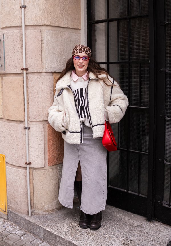 Žena u šal s leopard uzorkom na glavi, crvenim naočalama, bijelom mekanoj jakni, grafičkom džemperu, sivoj suknji s prednjim prorezom, crnim čizmama i crvenom torbom stoji uz kameni zid.