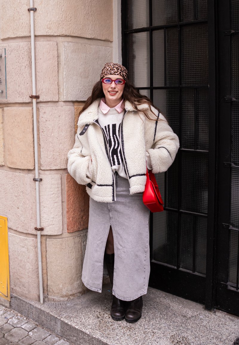 Kvinde med leopardmønstret tørklæde, røde briller, hvidt fluffy jakke, grafisk sweater, grå nederdel med frontslids, sorte støvler og rød håndtaske står ved en stenmur.