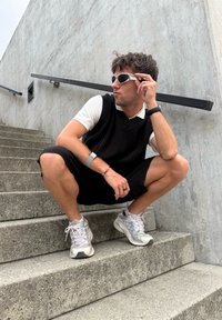 Pulôver preto de malha sem mangas sobre uma camisa pólo branca, calções pretos e ténis brancos gastos. Acessorizado com uma pulseira de prata e óculos de sol.