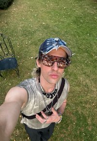Person wearing a distressed denim cap, oversized brown sunglasses, a textured grey sleeveless top, and layered silver and black necklaces. Grass background.