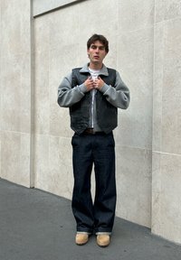 Sort lædervest over en grå sweatshirt, parret med mørkeblå brede jeans og beige støvler. Lige holdning mod en lys væg.