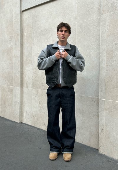 Chaleco de cuero negro sobre un sudadero gris, combinado con unos pantalones vaqueros de pierna ancha azul oscuro y botas beige. Postura recta frente a una pared clara.