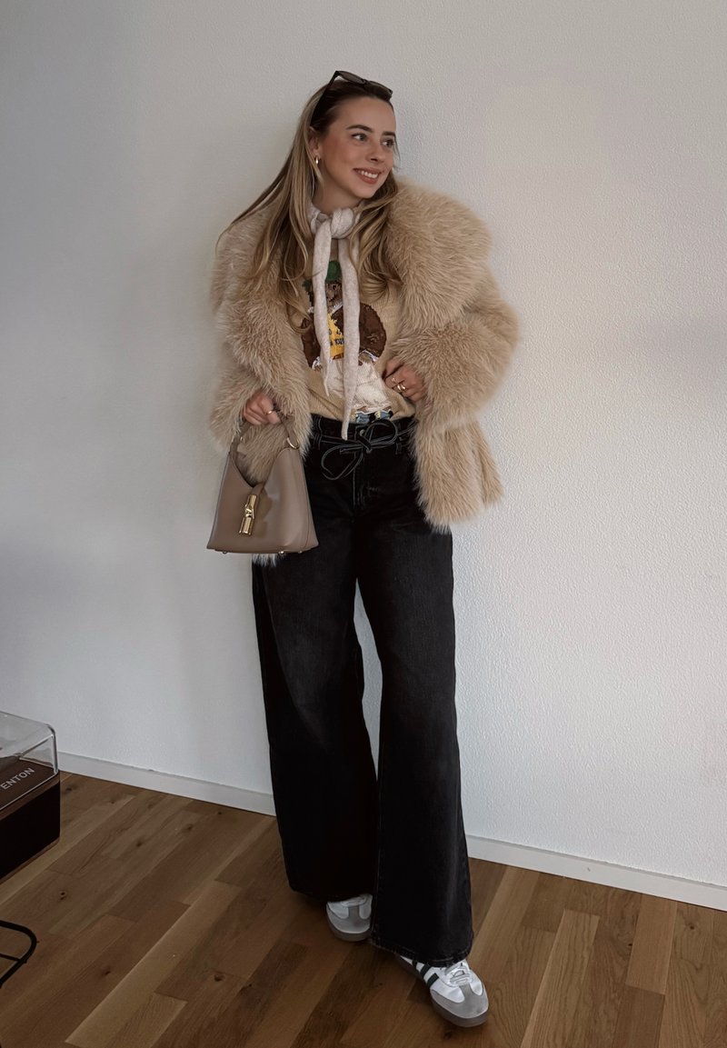 Manteau en fausse fourrure beige, pull graphique, pantalon noir à jambes larges et baskets blanches. Sac à main beige avec détails métalliques dorés. Fond en parquet en bois.