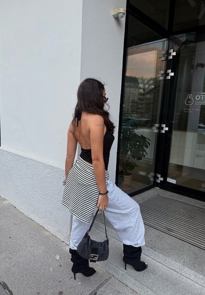 Top negro sin espalda, falda envolvente a rayas, pantalones sueltos grises, botas de tacón negras y un pequeño bolso negro, de pie junto a una puerta de cristal.