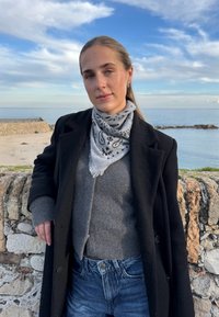 Jeune femme portant un manteau noir, un pull gris et une écharpe à motifs, debout contre un mur de pierre avec la mer calme et un ciel bleu en arrière-plan.