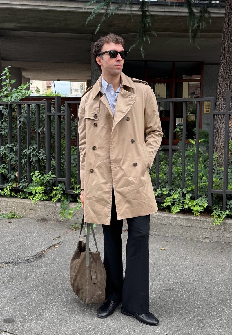 Homemem com um trench coat bege, óculos de sol pretos, calças e sapatos pretos, segura uma bolsa de tote castanha enquanto está de pé num passeio urbano perto de um edifício e de vegetação.