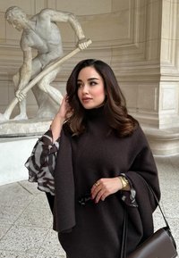 Ruskea kangaspäiväpeite, jossa on korkea kaulus ja läpikuultavat kuviolliset hihat. Pitelee aurinkolaseja, asusteena kultakello ja sormus.