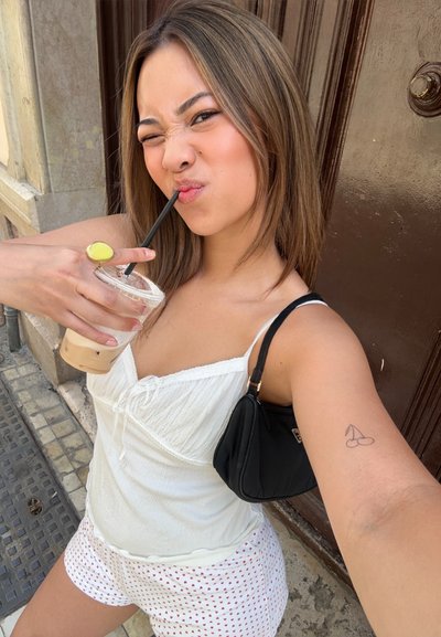 Camiseta blanca sin mangas con detalles de lazo, pantalones cortos estampados con puntos rojos y un bolso negro; sosteniendo un café helado con una pajita negra.