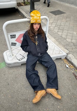 Veste en denim foncé et pantalon ample. Bonnet jaune avec logo. Bottes marron avec semelles crantées. Lunettes sur le sol à côté.