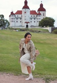 Beige trench coat worn over a white lace dress, paired with white block-heeled boots and a small tan woven handbag, set against a historic building and green grass.