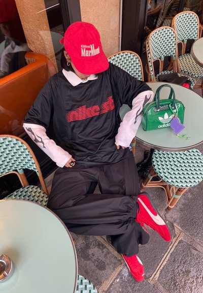 Persona que lleva una gorra roja de "Château Marlmont", camiseta negra de Nike, mangas largas blancas, pantalones negros y zapatillas rojas, sentado en una mesa al aire libre de un café.