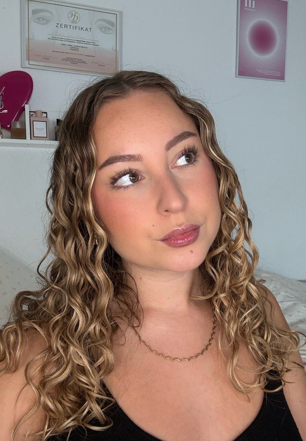 Jeune femme aux cheveux blonds et bouclés, portant un haut noir et un collier en or, regarde légèrement vers le haut avec une expression pensive.