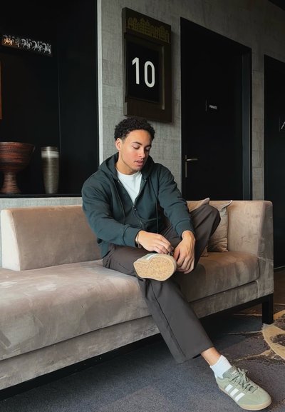 Sudadera gris con cremallera, camisa blanca, pantalones oscuros y zapatos verdes con acentos blancos. Sentado en un sofá ligero de terciopelo gris en un espacio interior.