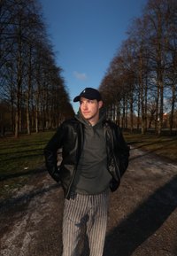 Crna kožna jakna preko sive dukserice, prugaste hlače i mornarsko plava kapa. Na otvorenom, s drvenom stazom i plavim nebom u pozadini.