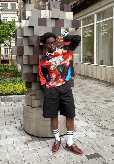 Camisa de mangas largas colorida con espirales rojas, azules y negras, pantalones cortos negros, calcetines blancos con rayas negras y zapatos marrones. Arte geométrico al aire libre de fondo.