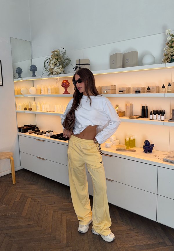 Frau mit Sonnenbrille, weißem, kurz geschnittenem Langarmshirt und gelber Hose steht in einem modernen Geschäft mit Regalen voller Hautpflegeprodukte und Dekor.