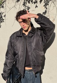 Melns neilona bumbereiņu jakas ar rāvējslēdzēju priekšpusē, lielām kabatām un ribinātu apkakli, uzvilkta virs melnas kreklas un apvienota ar džinsiem.