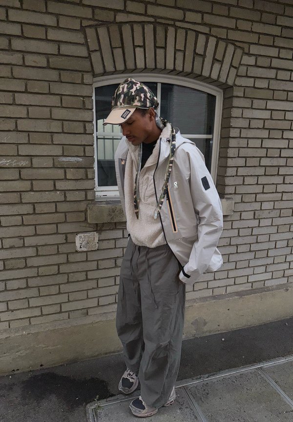 Lichtgrijze jas met rits en reflecterende accenten, gedragen over een beige gestructureerde fleece en grote, losvallende grijze broek. Camouflagepet.