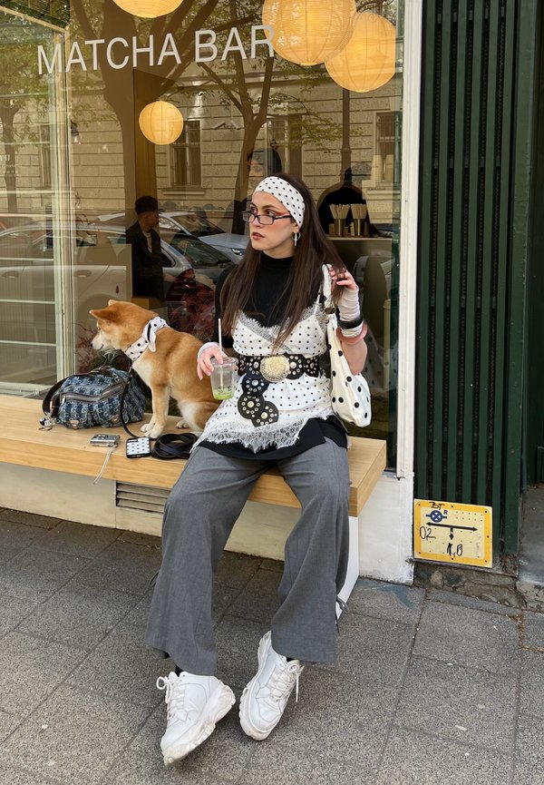 Γυναίκα με ρούχα που έχουν πουά σχέδιο και κορδέλα στα μαλλιά κάθεται σε παγκάκι έξω από το Matcha Bar, κρατώντας ένα ποτό, με έναν σκύλο Shiba Inu να φοράει το ίδιο φουλάρι δίπλα της.