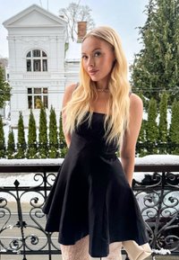 Femme aux cheveux longs et blonds portant une robe noire et des collants en dentelle, debout sur un balcon enneigé avec une maison blanche et des arbres en arrière-plan.