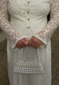 Ženska v beli čipkasti obleki drži biserno torbico z obema rokama pred pasom.