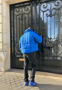 Lyseblå dunjakke, sorte jeans og blå sko. Personen rækker mod en dekorativ sort smedejernsdør.