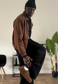 Casaco de lã castanho com uma camisa preta, calças pretas e sapatos em camurça castanha. Meias brancas visíveis. Planta em vaso ao fundo.