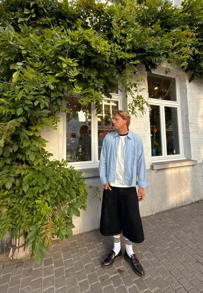 Camisa de cuadro azul claro, camiseta blanca, pantalones cortos negros hasta la rodilla, calcetines a rayas, zapatos negros; follaje y ventana en el fondo.