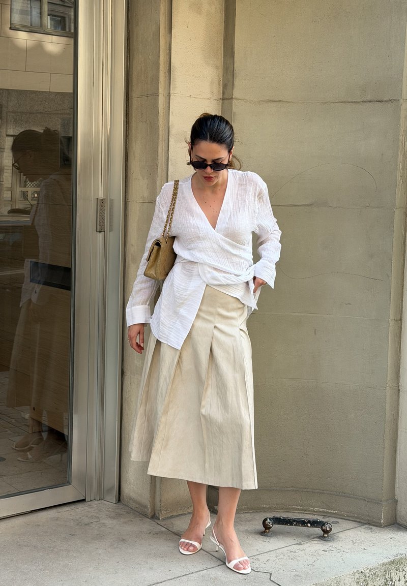 Frau mit Sonnenbrille, weißer Wickelbluse, beigem plissiertem Rock, weißen Sandalen und beiger Umhängetasche, die an einer Glastür und Steinmauer steht.