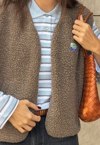 Brūns vilnas bezpiedurkņu žakets ar apaļu kakla izgriezumu, valkāts virs gaiši zilās svītrainās krekliņas. Apģērbs ietver austu oranžu rokassomu un brūnu jostu.