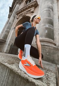 Brooks löparskor i klar orange med vita detaljer, med ett mesh-överdrag, tjock sula och logotyp. Bärs med svarta leggings och vita strumpor.