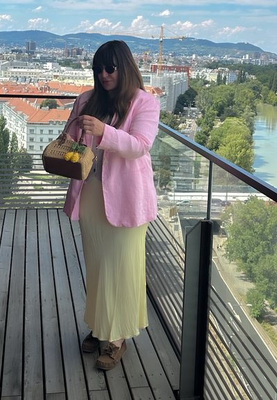 Blazer de lino rosa oversize, top blanco y falda maxi amarilla fluida. Sosteniendo un bolso de mimbre con acentos amarillos. Balcón de madera con vista a la ciudad.