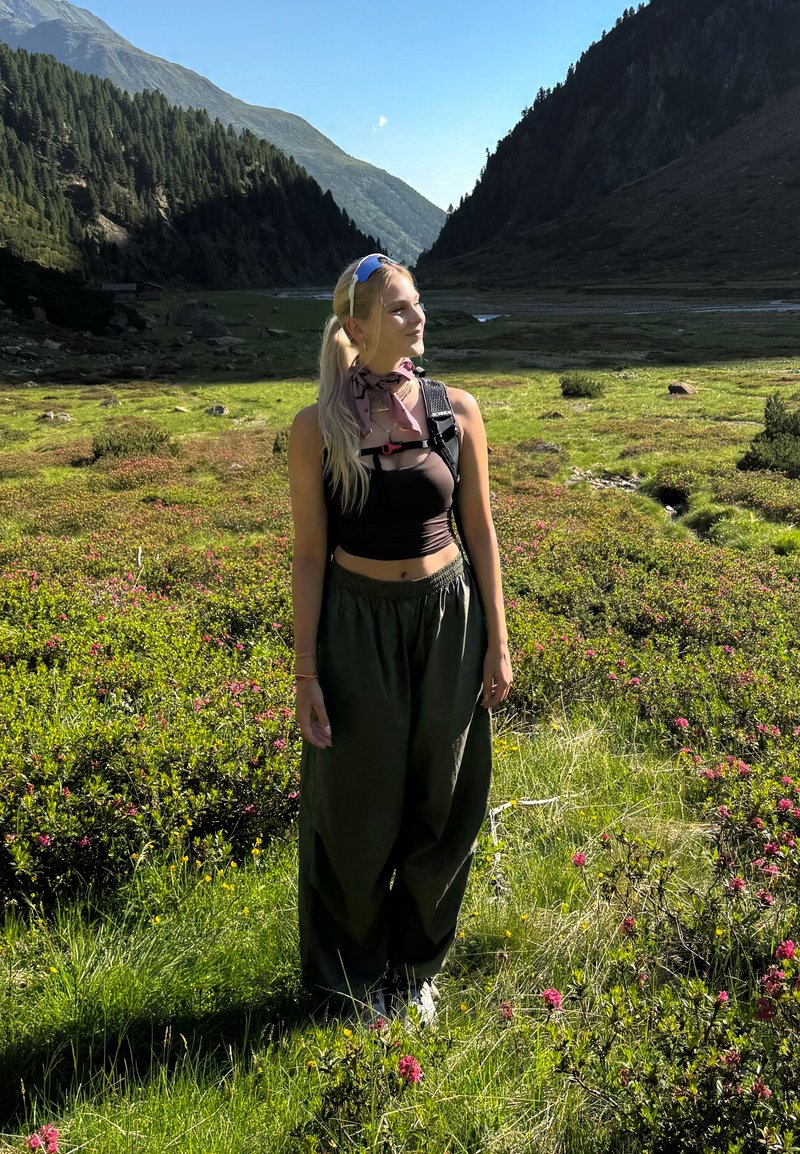 Jeune femme aux longs cheveux blonds debout dans une prairie de montagne verte avec des fleurs sauvages, portant un haut noir, un pantalon ample, un sac à dos et des lunettes de soleil.
