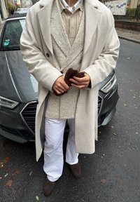 Lysegrå kåpe over en beige vest og kremfarget skjorte; hvite bukser og brune hansker. Står ved siden av en mørkegrå bil, på en våt gate.