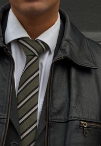 Jaqueta de couro preta sobre um colete cinza e uma camisa branca, com uma gravata listrada em verde e preto. Detalhe notável do fecho no casaco.