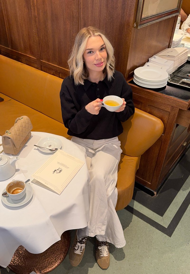 Femme aux cheveux blonds, pull noir et pantalon blanc assise sur un banc moutarde, tenant une tasse de thé à une table avec une théière et un livre.