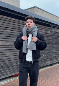 Écharpe en laine grise enroulée autour du cou d'une personne, veste bomber noire, pull en maille torsadée blanc et pantalon foncé devant un fond en bois.