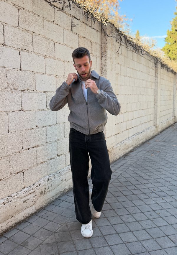 Homme debout sur un trottoir carrelé près d'un mur de pierre, ajustant le col d'une veste grise, portant un pantalon noir et des baskets blanches.