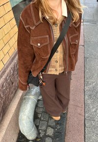 Hnědá semišová bunda s bílým prošíváním, pod ní khaki svetr na knoflíky, hnědé kalhoty s proužky a černá crossbody taška s přívěsky na klíče.